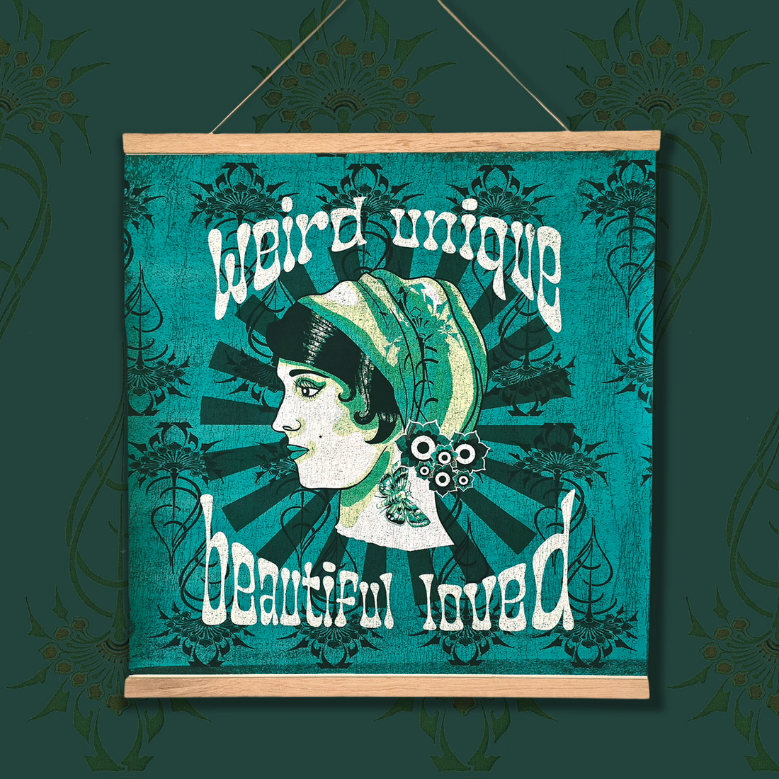 A square hanging wall art print with a light wood top and bottom frame, featuring a stylized profile illustration of a woman in a green and cream headscarf on a teal background with decorative text reading 'weird unique beautiful loved.' The piece is shown against a dark teal patterned backdrop.